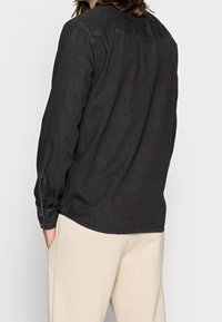 Czarna koszula dżinsowa z długimi rękawami, mankietami na guziki i detalem w kształcie joki na plecach. Posiada luźny krój i delikatny połysk.