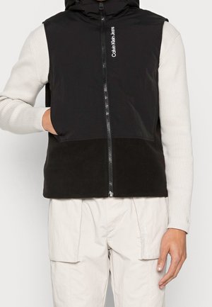 Person iført en ærmeløs sort vest med lynlås og teksten "Calvin Klein Jeans", cremefarvet ribbet langærmet skjorte og lyse cargobukser.