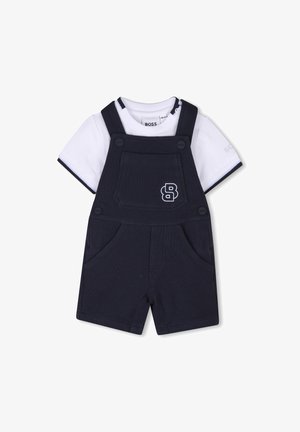 Salopette en coton bleu marine avec bretelles, dotée d'une poche avant et de poches latérales. Le t-shirt blanc en dessous a un passepoil bleu marine et un logo.