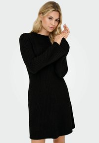 Frau mit blonden Haaren, die ein langärmliges schwarzes Rippstrickkleid trägt und mit den Händen nahe dem Gesicht vor einem schlichtem hellen Hintergrund posiert.