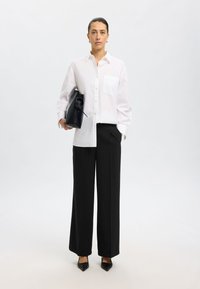 Witte button-up shirt met een zak, gecombineerd met hoge zwarte wijde broek en zwarte puntige hakken, met een zwarte tas.
