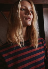 Triibuline pikkade varrukatega t-särk, millel on punased ja mereväesinised horisontaalsed triibud. Pehme kangas, lõdvestunud istuvus ja ümmargune kaelus. Looduslik juuksed, mis raamivad nägu.