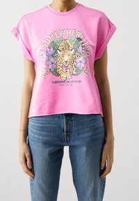 Mujer vestida con una camiseta de manga enrollada rosa con un diseño de leopardo y flores y el texto "Escapa a un lugar tropical" y vaqueros azules.