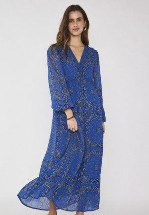 Donna che indossa un lungo abito blu con motivi paisley, maniche lunghe e scollo a V, in piedi davanti a uno sfondo chiaro uniforme.
