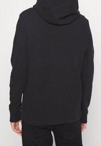 Sudadera negra con un tejido texturizado, que cuenta con una gran capucha y mangas largas. La parte posterior tiene un acabado liso sin detalles adicionales.