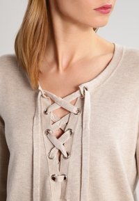 Beige knit sweater featuring a lace-up front with silver grommets. Round neckline and a soft texture complete the design.