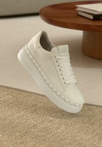 Άσπρο δερμάτινο αθλητικό παπούτσι με υφή στην επιφάνεια, επίπεδα κορδόνια και διακοσμητικά καρφιά κατά μήκος της σόλας. Στρογγυλερή μύτη και μίνιμαλ σχεδιασμός.