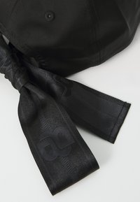 Berretto nero con finitura in tessuto liscio, caratterizzato da una corona alta e due lunghe cordicelle intrecciate che mostrano un motivo logo delicato.