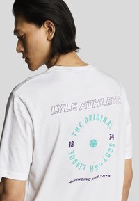 Hvid bomulds-T-shirt med lilla og teal tekst. Designet inkluderer "LYLE ATHLETIC," "THE ORIGINAL SCOTTISH LEAGUE," og et fodboldgrafik.