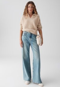 Beige pluizig sweater met een V-hals, gecombineerd met lichtblauwe wijde jeans en witte instappers. Houdt een kleine beige tas vast.