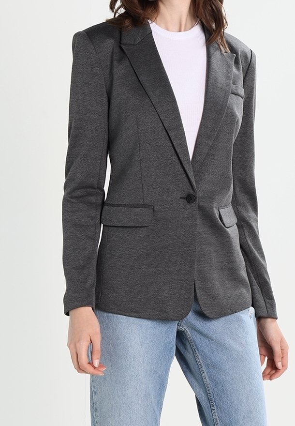 Femme portant un blazer gris foncé ajusté sur une chemise blanche, assorti à un jean en denim bleu clair sur un fond uni.