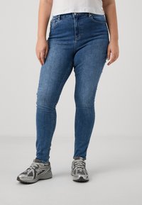 Λεπτά μπλε τζιν από denim, με ψηλή μέση, διακριτική φθορά και πίσω τσέπες. Συνδυασμένα με γκρι αθλητικά παπούτσια.