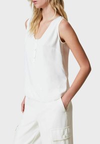 Top bianco senza maniche con scollatura a V, dettaglio a bottoni sul davanti e bordi smerlati. Tessuto morbido, abbinato a pantaloni bianchi con tasche.
