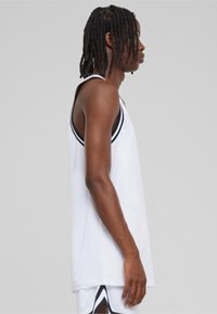 Hvit mesh tank topp med svart piping, med en løs passform og brede armhull, vist fra siden for å fremheve det lette designet.
