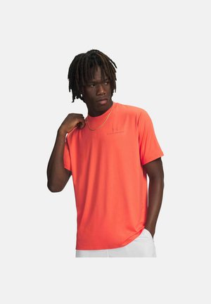 Uomo con dreadlocks che indossa una maglietta sportiva a maniche corte di un arancione brillante e pantaloni bianchi, che tocca il colletto della maglietta con una mano.