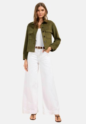 Žena v olivově zelené zapínané bundě přes bílý top, s vysoce pasovanými širokými bílými kalhotami, hnědým páskem a hnědými sandály stojí na bílém pozadí.