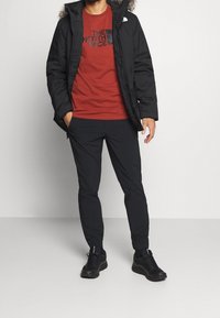 Veste isolée noire avec capuche doublée en fourrure, t-shirt rouge avec logo noir, pantalon noir et chaussures de sport noires.