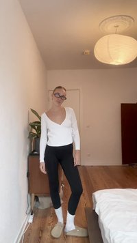 Weißes langärmliges Wickeloberteil mit tiefem V-Ausschnitt, kombiniert mit schwarzen Leggings und beigen Hausschuhen. Schlichte Gestaltung, weicher Stoff, minimale Details.