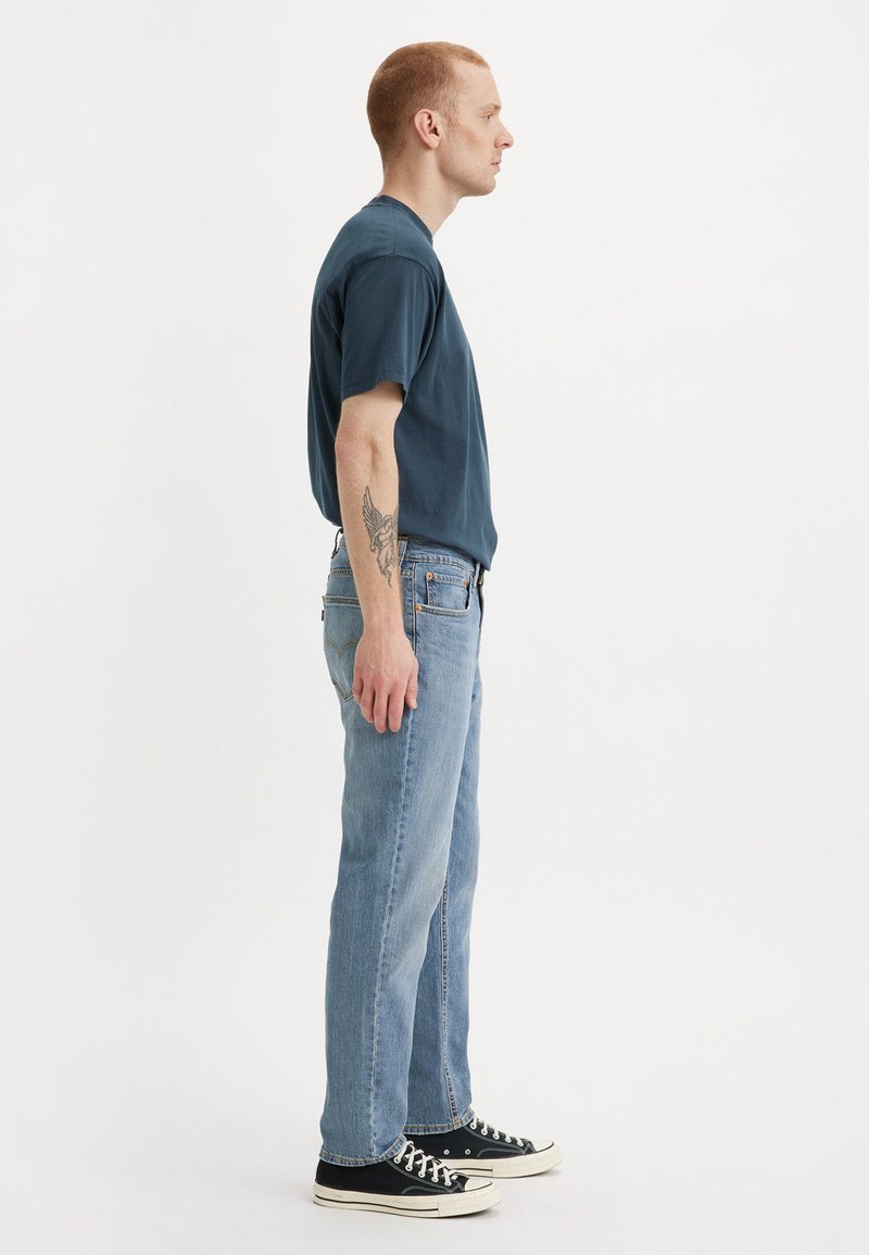 T-shirt à manches courtes bleu foncé, jean bleu clair, baskets noires avec semelles blanches, et tatouage visible sur le bras droit. Vue de profil.