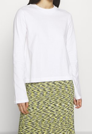 Femme portant une chemise blanche unie à manches longues et une jupe à motifs jaune, gris et noir sur fond blanc.