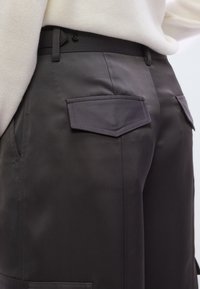 Tmavosivé nohavice z hladkej látky s dvoma zadnými vreckami so špicatými chlopňami a priliehavým pásom. Minimálne viditeľné švy.