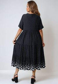 Zwarte jurk met korte mouwen, witte stippen, gelaagd ontwerp en een zoom met grotere witte stippen. Platte zwarte hakken.
