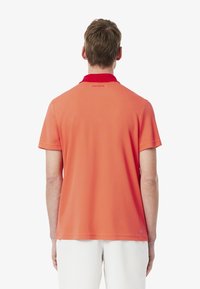 Pikétröja i stark korall med en kontrasterande röd krage. Har en liten präglad logotyp på baksidan och en mjuk, lätt tygkvalitet.