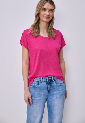 Vrouw met steil blond haar, gekleed in een felroze T-shirt met korte mouwen en lichtblauwe spijkerbroek, staand tegen een eenvoudige lichte achtergrond.