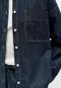 Donkerblauw denim overhemd met knopen, voorzien van een borstzak, witte knopen en contrasterende stiksels. Casual ontwerp, zachte textuur.