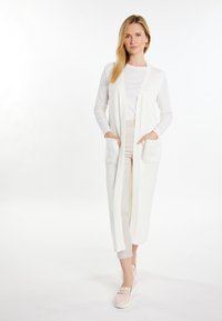 Cardigan lung alb fabricat din material ribat, cu două buzunare frontale, asortat cu o bluză ușoară și pantaloni bej, având o croială lejeră.