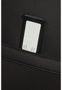 Porte-étiquette en tissu noir avec une étiquette blanche affichant des lignes pour le nom, l'adresse, le téléphone et l'email, accrochée à un fond texturé.