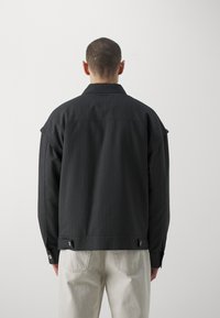 Svart randig jacka med en avslappnad passform, en spetskrage och metallknappar vid fållen. Återfinns i kombination med ljusa, strukturerade byxor.