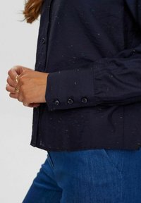 Mørkeblå skjorte med knapper og farverige prikker, lange ærmer og knaplukning ved håndleddene, parret med blå denim. Blødt stof og løs pasform.