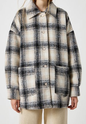 Femme portant une veste en laine beige et noire à carreaux oversize avec des boutons à l'avant et de grandes poches plaquées, associée à un pantalon clair.