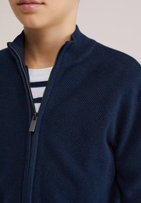 Tamsiai mėlynas užtraukiamas švarkas su faktūruotu megztu raštu ir aukštu apykakliu. Pagamintas su sidabriniu užtrauktuku ir po juo baltas dryžuotas marškinėlis.