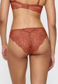 Koronkowe majtki w kolorze rdzy z kwiatowym wzorem, z falbanką na krawędzi i gładkim paskiem w talii. Miękka, przezroczysta tekstura koronki.
