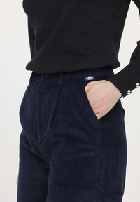 Pantalons en velours côtelé marine avec une taille haute, des poches latérales et une petite étiquette logo. Le tissu présente une texture côtelée, ajoutant un intérêt visuel.
