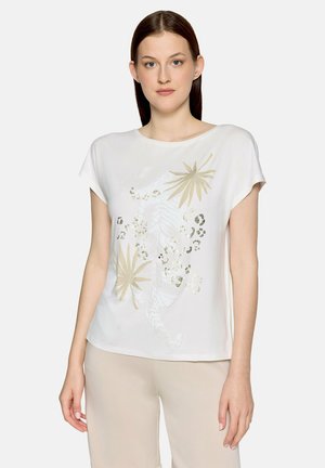 Jeune femme portant un haut blanc à manches courtes avec un motif de feuilles tropicales beiges et un imprimé léopard, associé à un pantalon beige, debout devant un fond blanc.