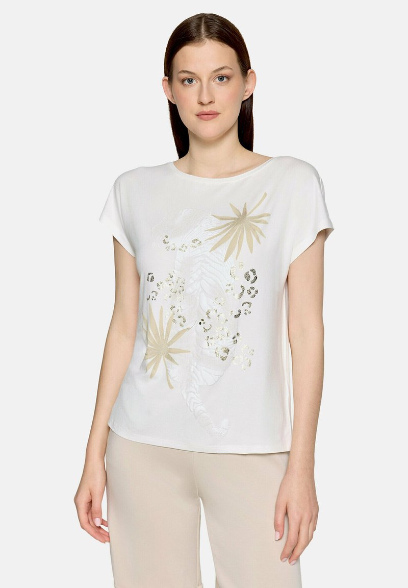 Jeune femme portant un haut blanc à manches courtes avec un motif de feuilles tropicales beiges et un imprimé léopard, associé à un pantalon beige, debout devant un fond blanc.