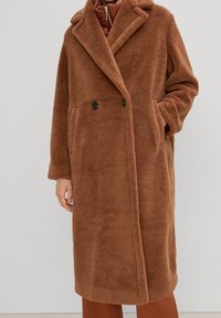 Abrigo teddy largo marrón con amplios solapas, cierre de doble botón y bolsillos laterales, llevado sobre un conjunto color óxido.