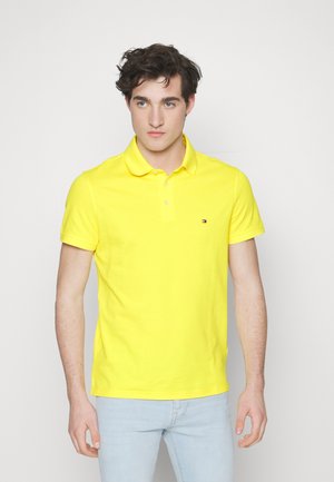 Polo giallo realizzato in cotone, con colletto bottonato, maniche corte e un piccolo logo sul lato sinistro del petto. Indossato con jeans azzurri chiari.