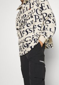 Sudadera de forro polar en crema con un patrón de texto en azul marino, cierre de media cremallera y puños bicolores. Combinada con pantalones cargo negros que tienen bolsillos laterales.