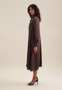 Lungo vestito fluido in tessuto scuro con motivo floreale, caratterizzato da maniche lunghe, un colletto e sandali neri con tacco alto.