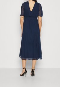 Mörkblå midi-klänning med V-rygg, korta ärmar och figursydd midja. Tyget är lätt och flödande, i kombination med svarta sandaletter med öppen tå.