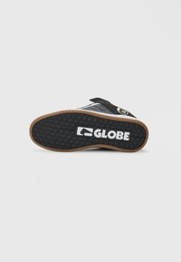Sole della sneaker Globe nera e marrone con battistrada testurizzato e logo Globe bianco centrato sulla suola, scarpa inclinata di lato su uno sfondo bianco.