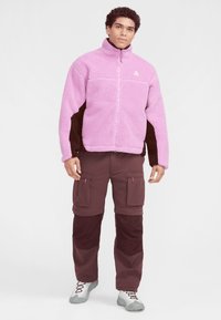 Fleecejacket i rosa med svarta sidopaneler, dragkedja fram, hög krage. Matcher med bruna cargobyxor med flera fickor.