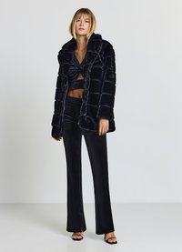 Cappotto in ecopelliccia nero con design trapuntato, colletto ampio, abbinato a un top corto nero e pantaloni in raso a vita alta, aderenti con spacchi laterali.