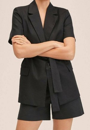 Personne portant un blazer noir à manches courtes et un short assorti, bras croisés, devant un fond beige uni.