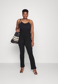 Top preto sem mangas com alças finas, calças pretas de cintura alta em malha e saltos altos pretos com tiras. Mão segurando uma bolsa tecida em preto e crème.