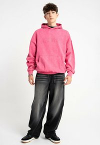 Różowa bluza z kapturem z kieszenią kangurką, z teksturowanym logo. Połączona z luźnymi czarnymi dżinsami i czarnymi trampkami z białymi podeszwami.